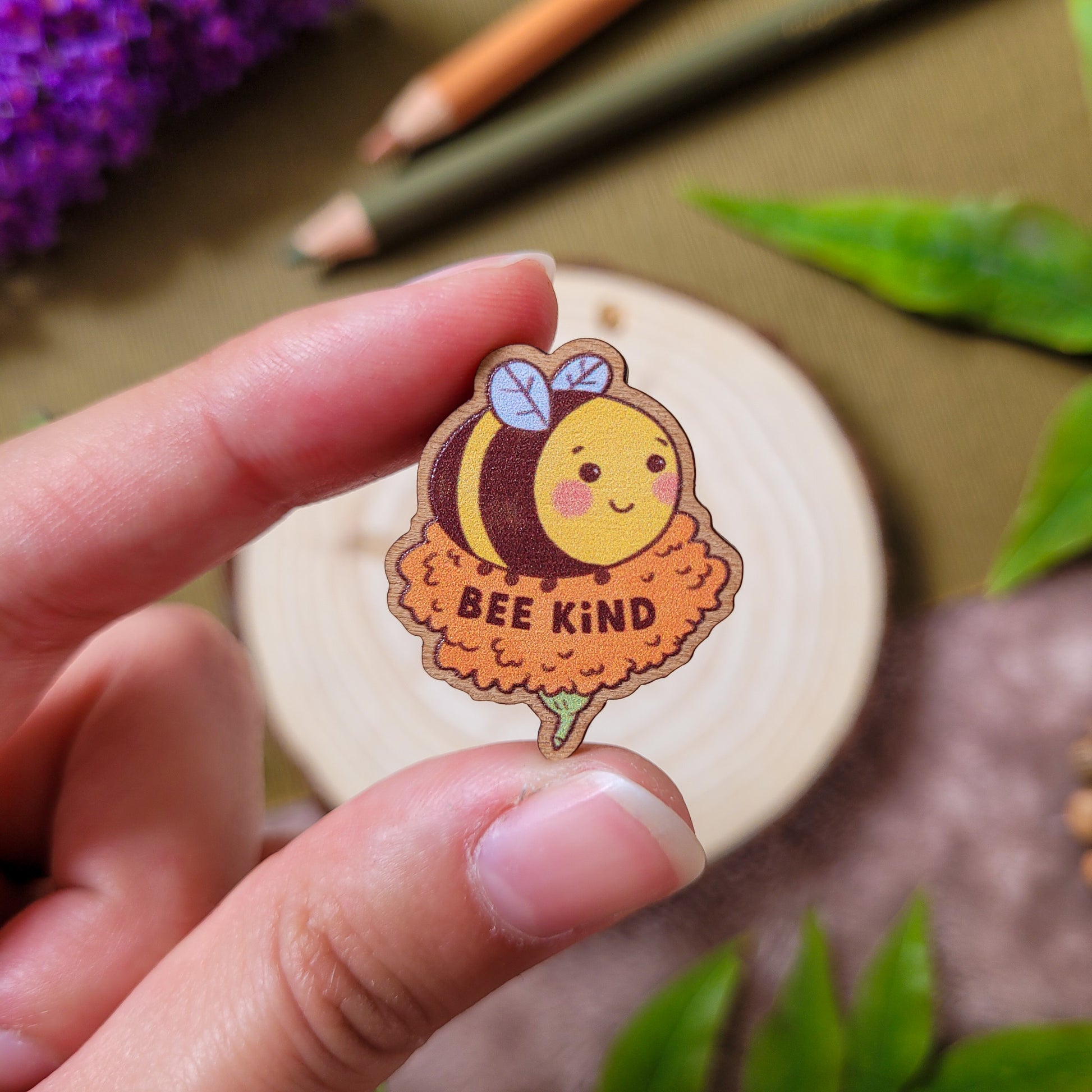 pins en bois d'une abeille mignonne posée sur une fleur orange avec un texte qui dit "Bee Kind". Fait à partir de bois éco responsable. Le pins est tenu dasn la main, il fait 3 x 2,5cm.
