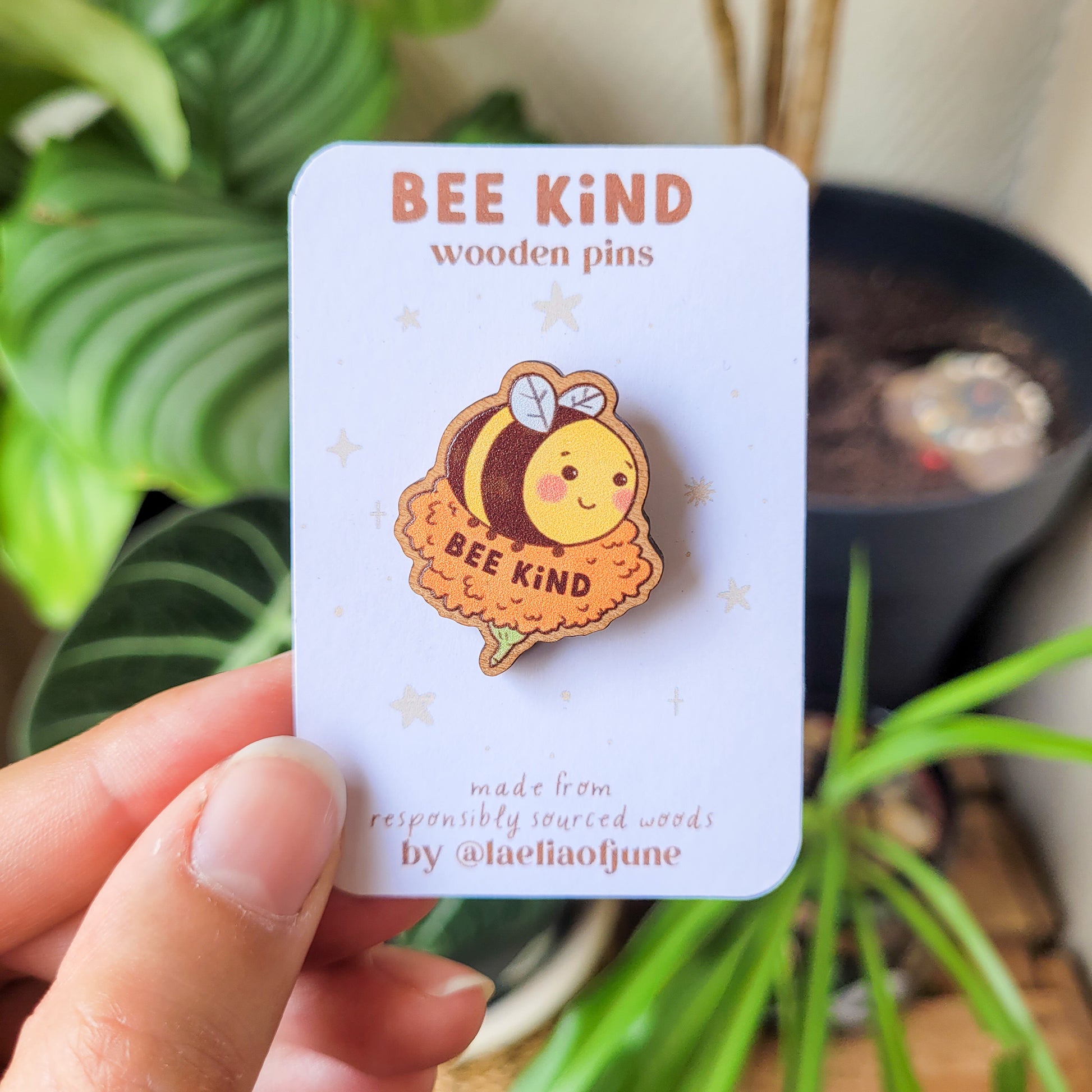 pins en bois d'une abeille mignonne posée sur une fleur orange avec un texte qui dit "Bee Kind". Fait à partir de bois éco responsable. Le pins est tenu dans la main avec son emballage.
