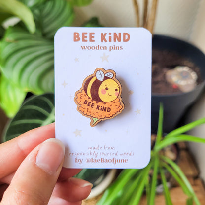 pins en bois d'une abeille mignonne posée sur une fleur orange avec un texte qui dit "Bee Kind". Fait à partir de bois éco responsable. Le pins est tenu dans la main avec son emballage.